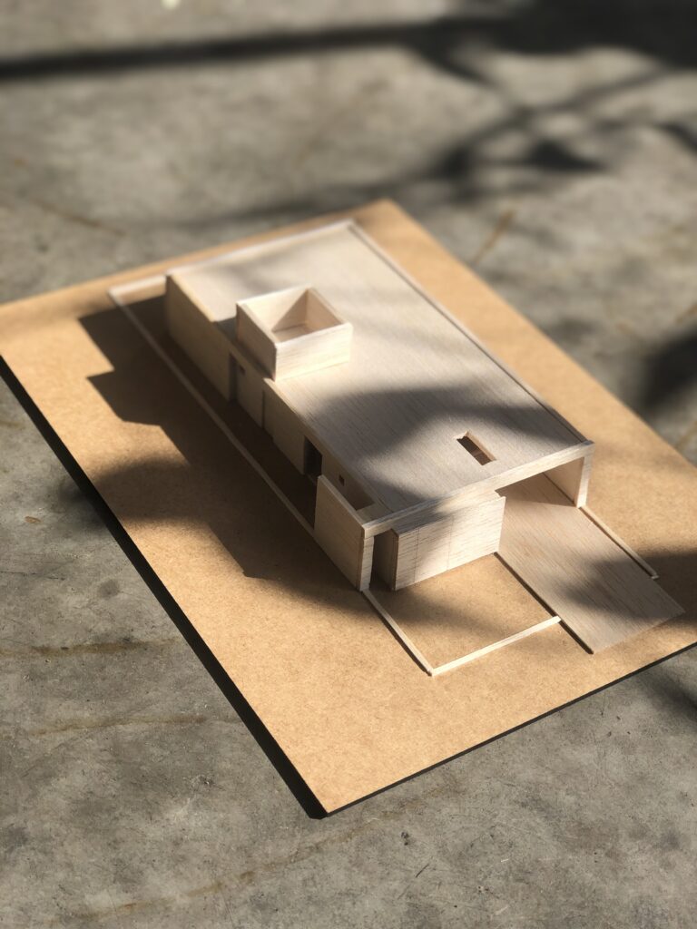 Imagem de uma maquete em madeira balsa, uma madeira bege clara, sobre uma base de madeira, sobre um piso de concreto cinza. A casa representada nessa miniatura é referente a etapa de projeto arquitetônico de estudo preliminar.