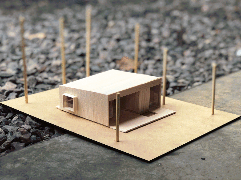 Imagem de uma maquete volumétrica em piso de concreto e brita. Maquete monocromática, em tom claro de madeira. Simula projeto da Casa Rio Novo