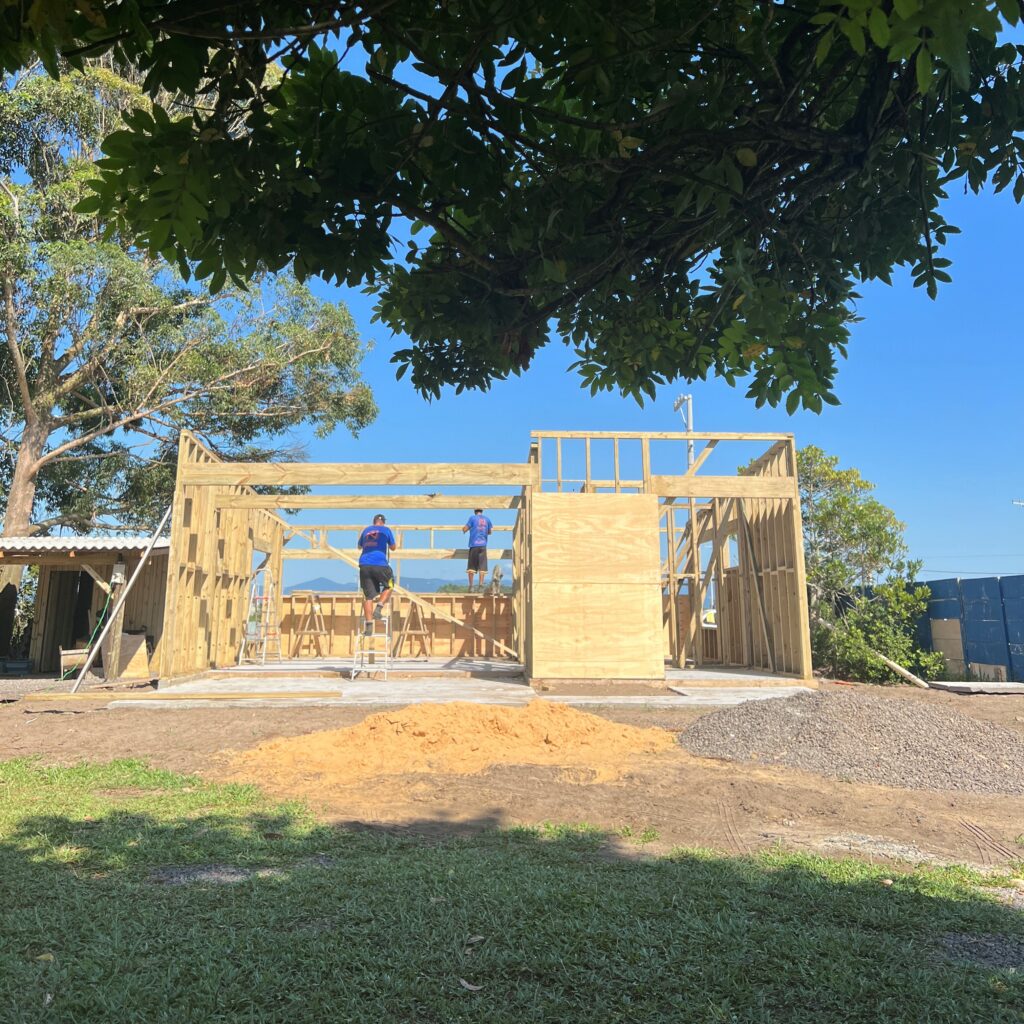 Foto de uma casa ainda em obras, estrutura de madeira e duas pessoas trabalhando na construção seca. Grama no entorno e céu azul, sem nuvens, con vegetação em primeiro plano funcionando como moldura da imagem