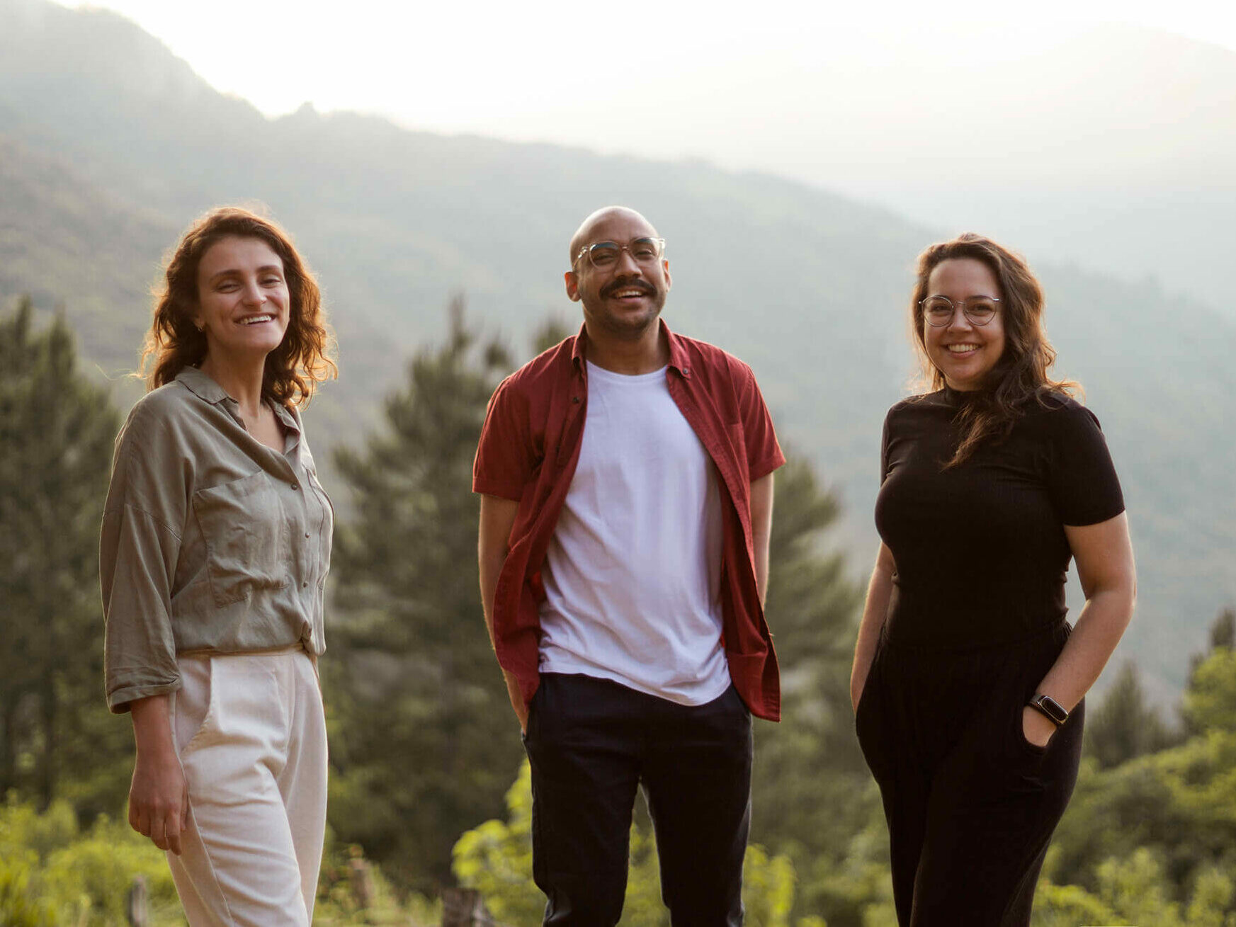 Foto de três pessoas, duas mulheres e um homem no meio, levemente separados, sorrindo, em primeiro plano, com vegetação ao fundo - os três arquitetos da Harpa esperando você entrar em contato para nos contratar