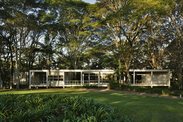 Imagem de uma casa com muita vegetação ao fundo. Casa em estilo de arquitetura atemporal, vê se duas lajes, a de piso e a de cobertura, na cor branca, conectada por colunas também brancas. A leveza da forma é destacada por grandes vidros e poucas paredes.